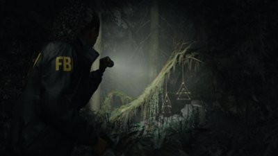 Captura de ecrã de Alan Wake 2 que mostra Saga Anderson com uma tocha a iluminar símbolos do oculto triangulares, pendurados num ramo de árvore na floresta