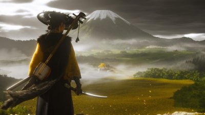 لقطة شاشة للعبة Ghost of Yotei تُظهر الشخصية الرئيسية تنظر إلى حقل من الأعشاب الذهبية، وجبل مغطى بالثلوج من بعيد