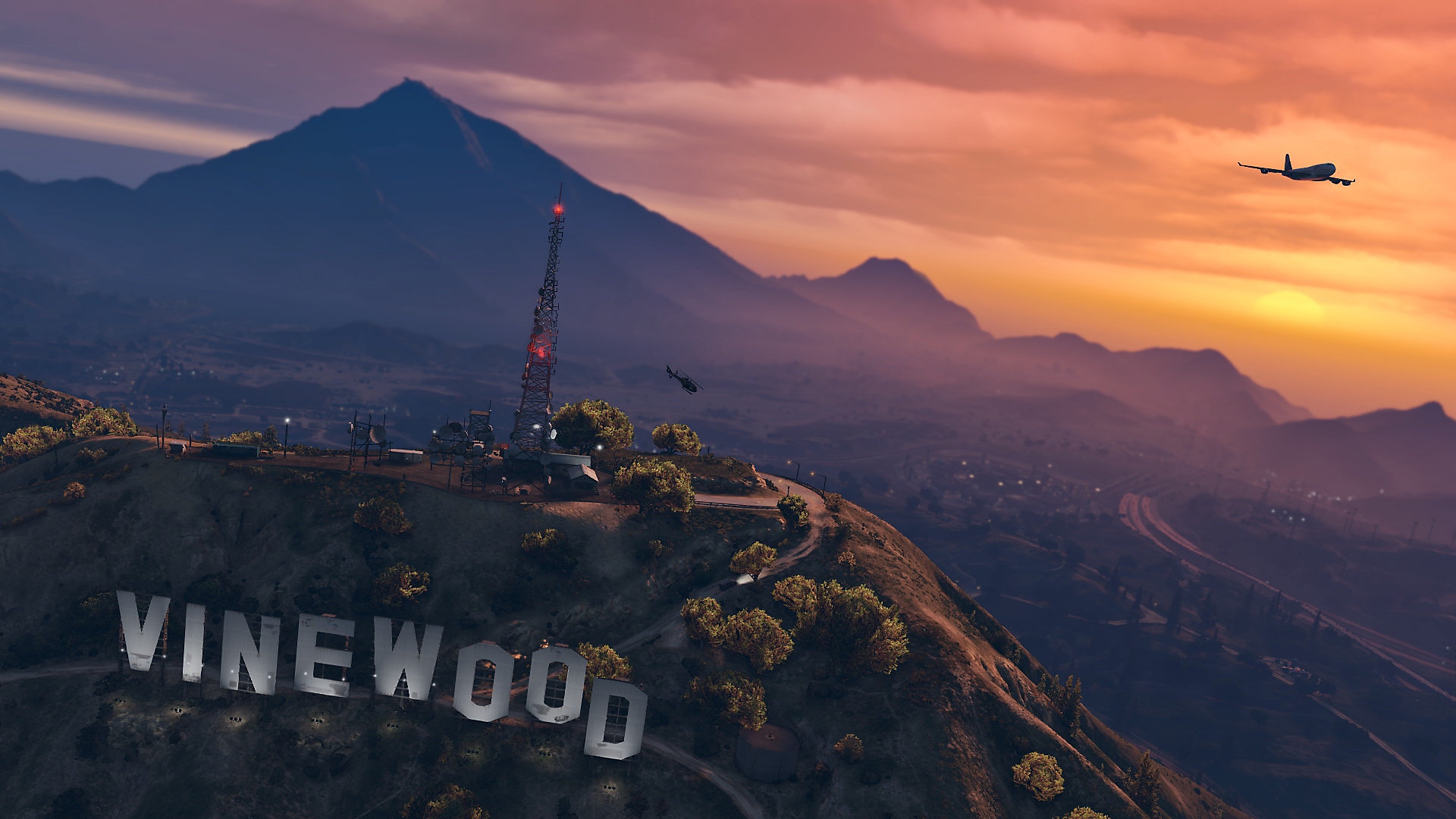 Grand Theft Auto V – snímka obrazovky zobrazujúca západ slnka nad kopcovitou krajinou s obrými písmenami vyvedeným nápisom „Vinewood“