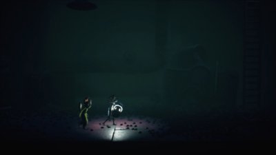 Ein Screenshot aus Little Nightmares 3, der Low zeigt, wie er eine Lichtquelle über einen dunklen Fabrikboden trägt 