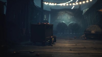 Ein Screenshot aus Little Nightmares 3, der zwei Gestalten zeigt, die gemeinsam eine Kiste über den Kirmesboden schleifen