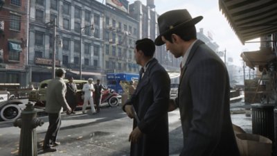Gameplay screenshot from Mafia: Definitive Edition depicting two men in early 20th century attire standing on a street corner.